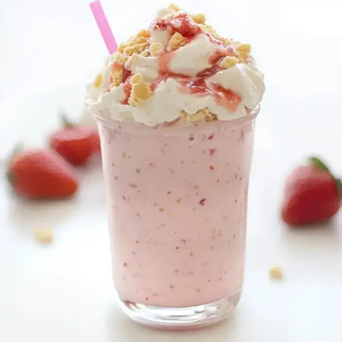 A milkshake in soft pink topped with whipped cream, cookie crumbles, and strawberry syrup, alongside fresh strawberries and a straw.