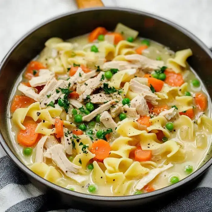 Close-up of a pot filled with creamy noodle soup featuring turkey pieces, egg noodles, carrots, and peas.