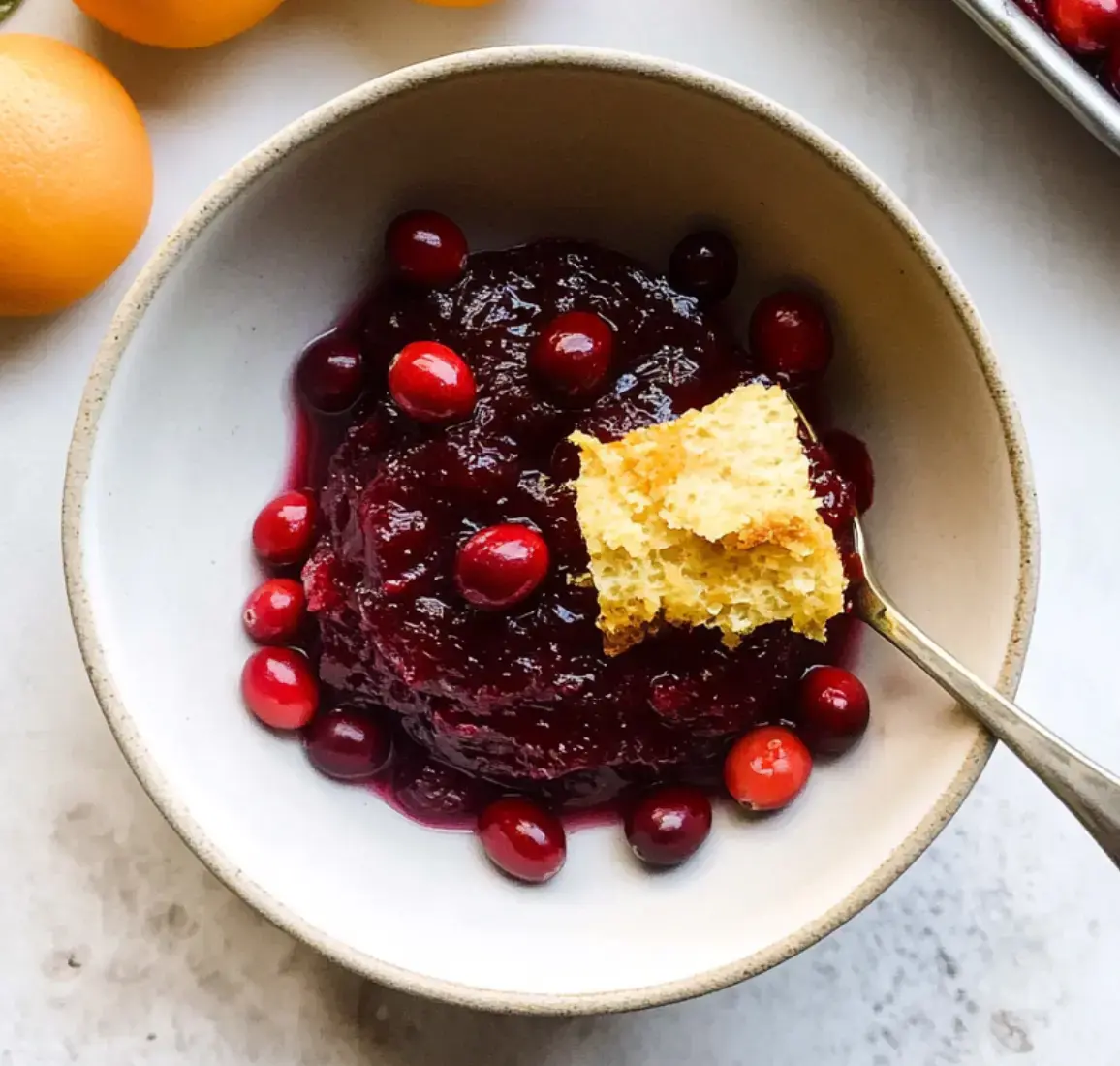A serving bowl of cranberry sauce with fresh fruit topping and cornbread on the side, set next to whole oranges.