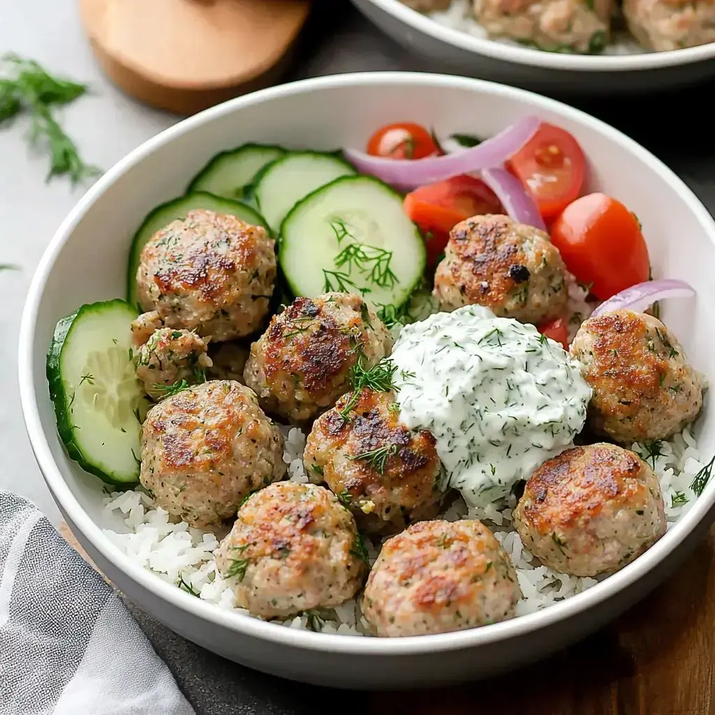 A bowl with rice, golden-brown meatballs, cucumber slices, cherry tomatoes, red onion, and a dollop of creamy sauce.