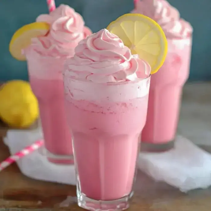 Three glasses filled with frothy pink drinks, cream on top, lemon slices, and straws, with a lemon on the side.