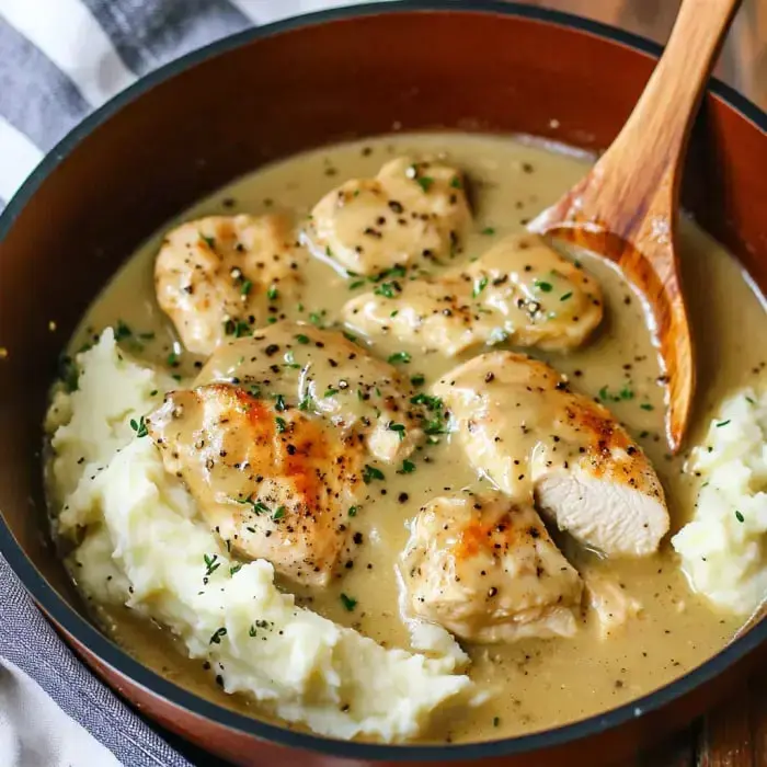 Bowl with creamy gravy-soaked chicken chunks on mashed potatoes, topped with fresh herbs.