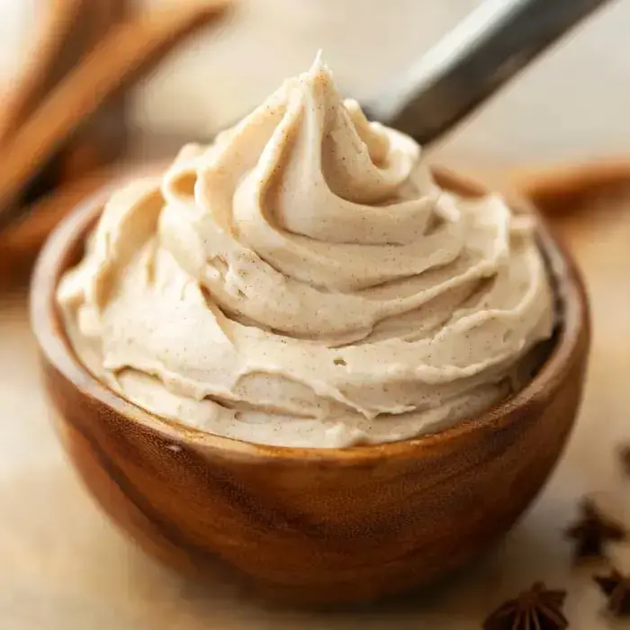A wooden bowl filled with creamy light brown frosting, swirled on top and surrounded by cinnamon sticks.