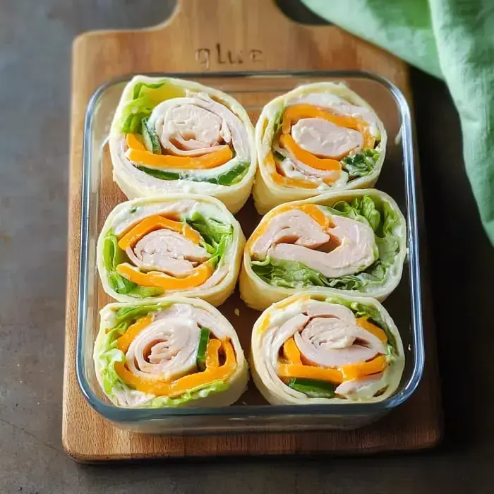 Six tortilla wraps with turkey, lettuce, and bright bell peppers line up neatly in a glass dish.