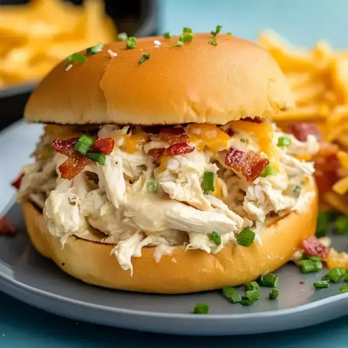A pile of shredded chicken topped with melting cheese, bacon, and green onions, tucked in a sandwich with fries on the side.