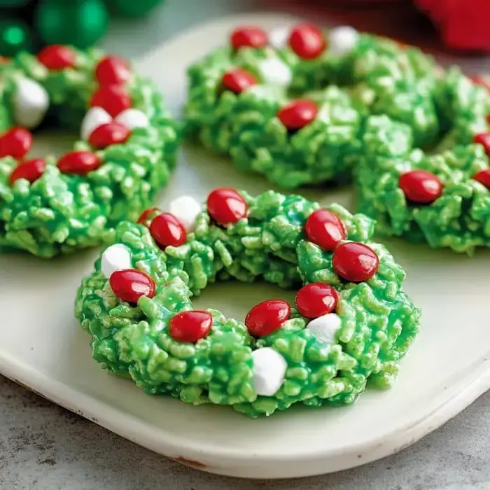 Festive green cereal wreath treats sitting on a plate, finished with white marshmallows and red candy decorations.