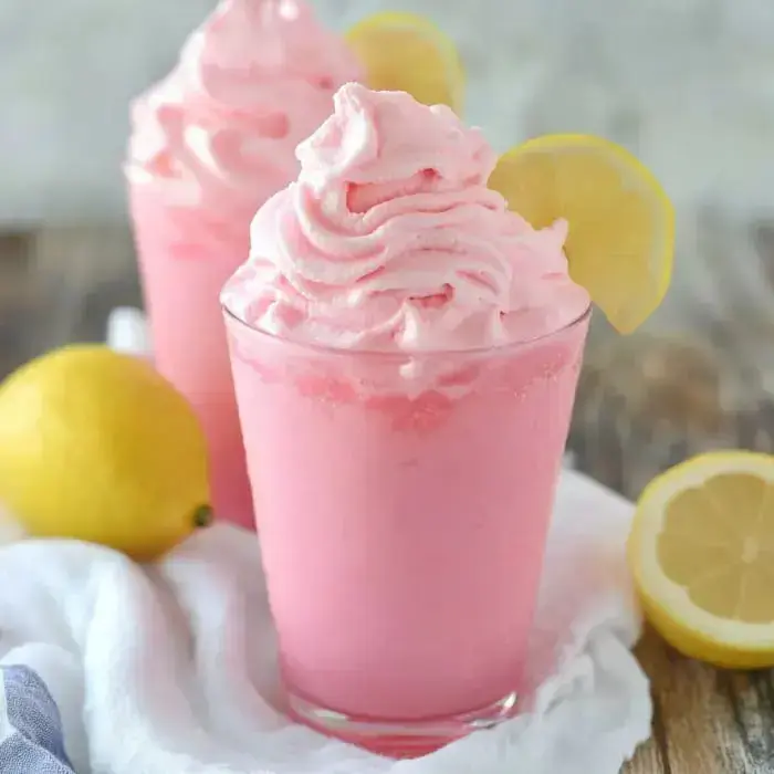 Glasses of frothy pink lemon drink with lemon wheels and fresh lemons sit on a soft cloth.