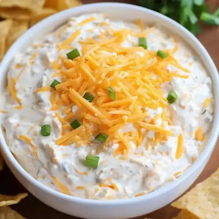 A bowl filled with creamy dip, fresh cheddar, and green onions sitting next to a pile of tortilla chips.