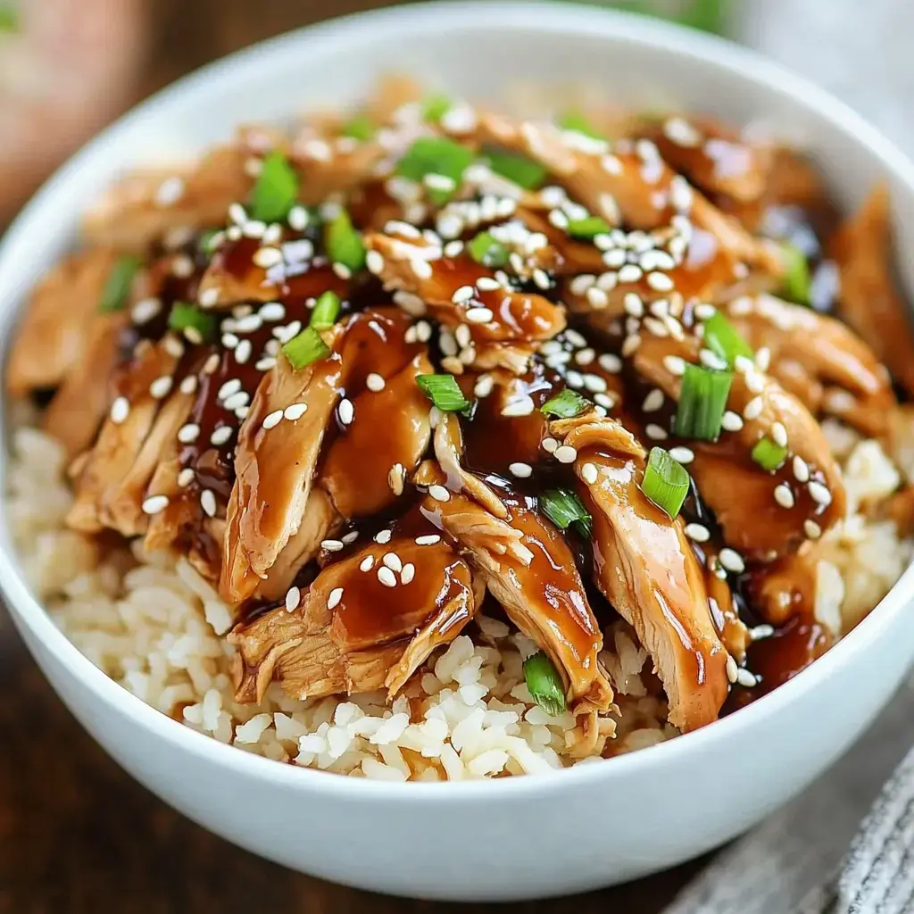 Closeup shot of rice in a bowl topped with sliced chicken, dark glaze, sesame seeds, and green onions.