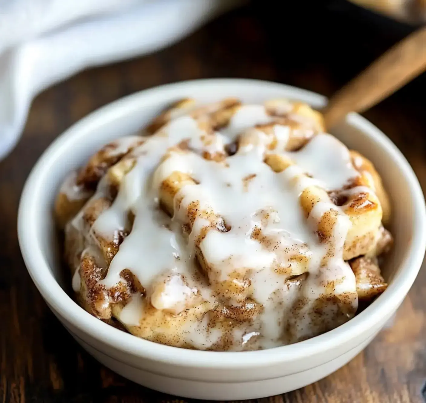 A bowl holding chunky cinnamon rolls with icing sits on a wood table.