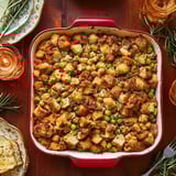 Golden baked stuffing paired with peas in a vibrant red dish on a cozy wooden table, surrounded by candles and festive plates.