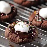 Chocolate cookies topped with melted chocolate, whipped cream, and sprinkles, cooling on a rack.