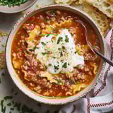 A cozy bowl filled with lasagna soup topped with ricotta cheese and herbs, served with crusty bread and fresh parsley.