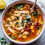 A bowl of shredded chicken soup with corn, diced tomatoes, cilantro, topped with green pepper pieces, and a lime wedge on the side.