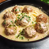 A dark bowl holding spaghetti with creamy sauce, topped with meatballs and fresh parsley.