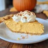 A creamy pumpkin pie slice topped with whipped cream and cinnamon, served on a white plate with a blurred pumpkin backdrop.