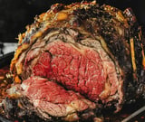 A juicy prime rib roast with a crispy herb crust, sliced to reveal its pink center and served on a dark plate.