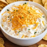 A bowl of creamy dip topped with shredded cheddar and herbs, surrounded by chips.
