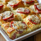 Crispy pastry squares stuffed with ham, cheese, and roasted peppers, sprinkled with herbs, displayed on a dish.